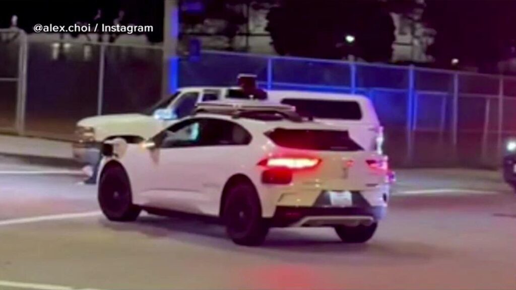 A driverless Waymo robotaxi at night with police emergency lights visible in the background