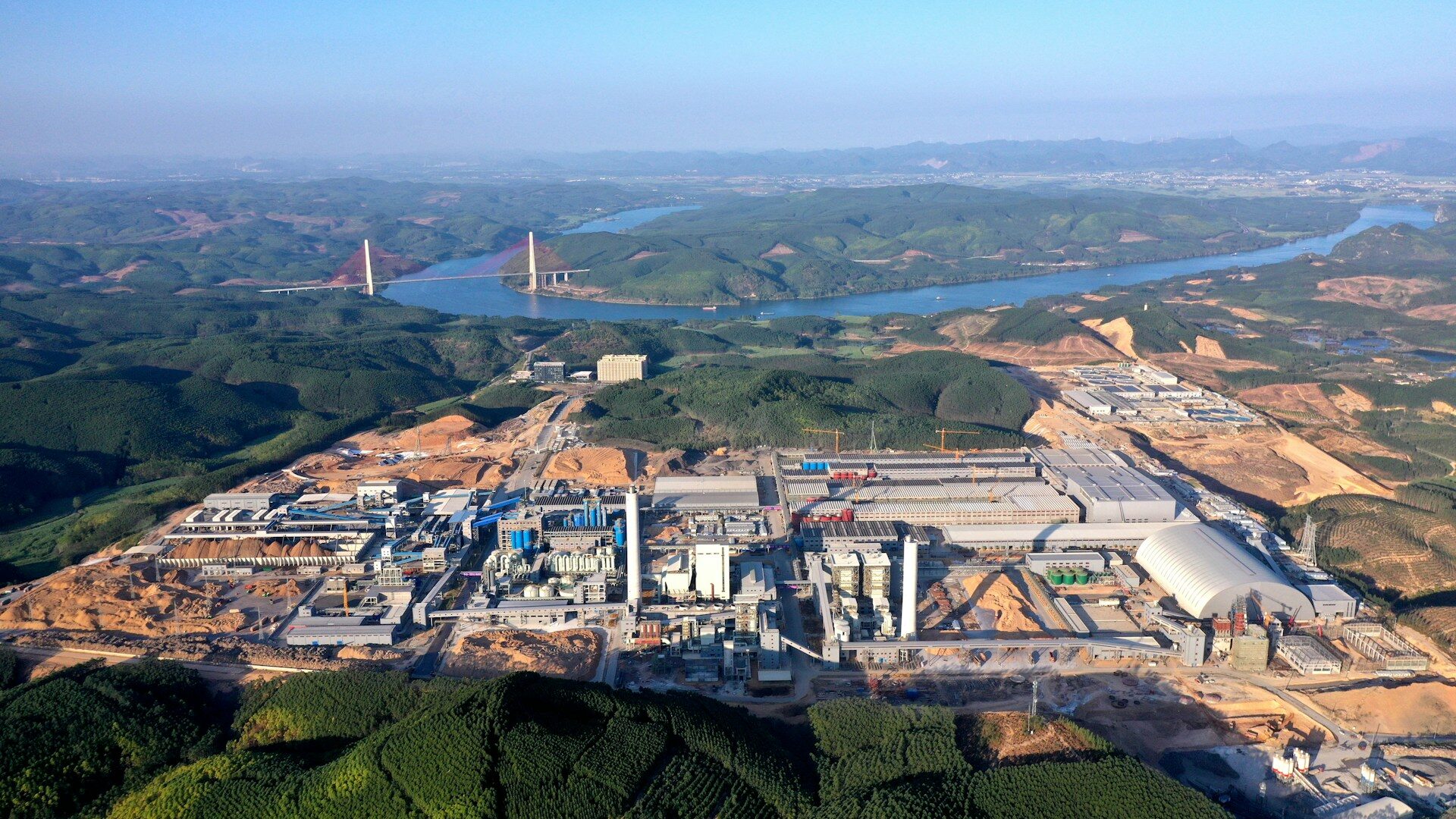 Industrial complex, green hills, river, and red suspension bridge.