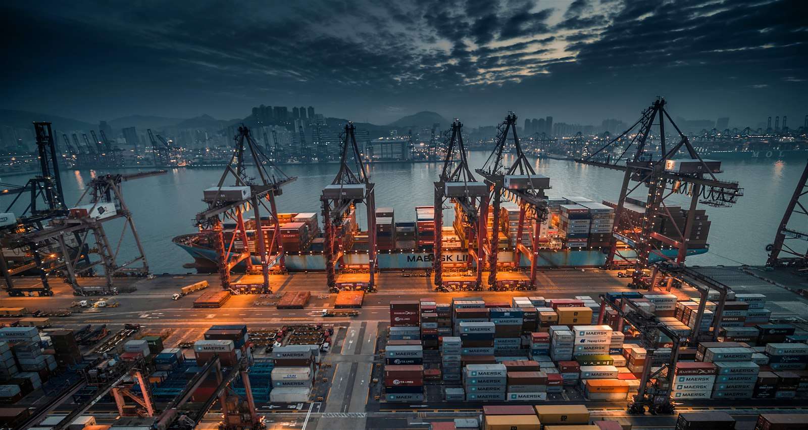 A container ship being loaded by cranes at an industrial port at dusk