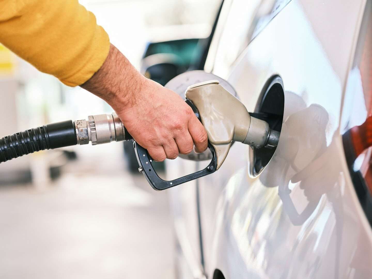 Driver refueling a white car at a gas pump, holding the nozzle in the filler neck