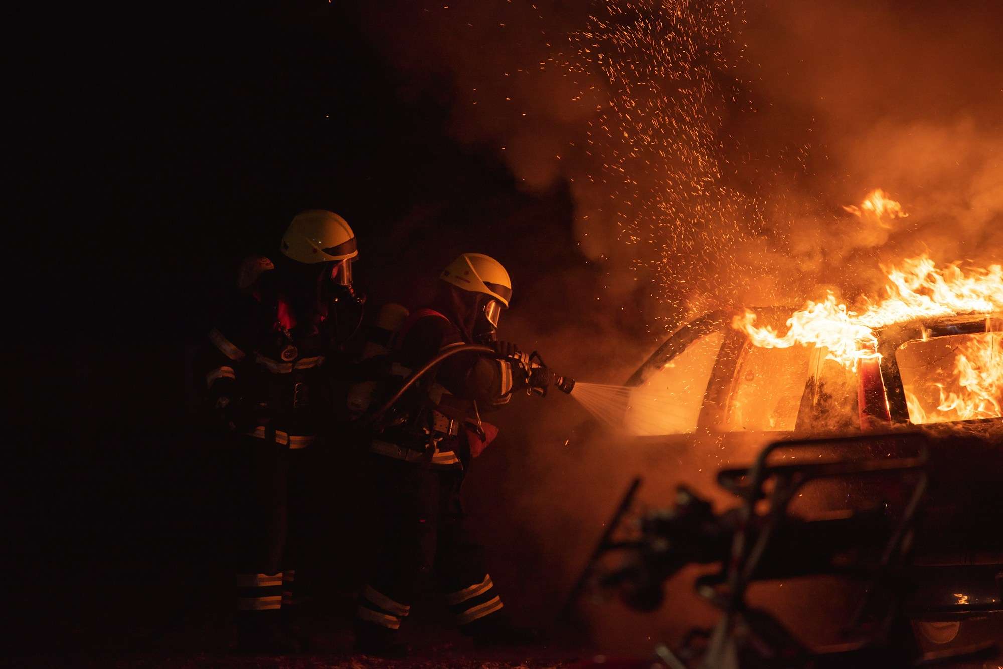 Two firefighters spray water on a burning car at night