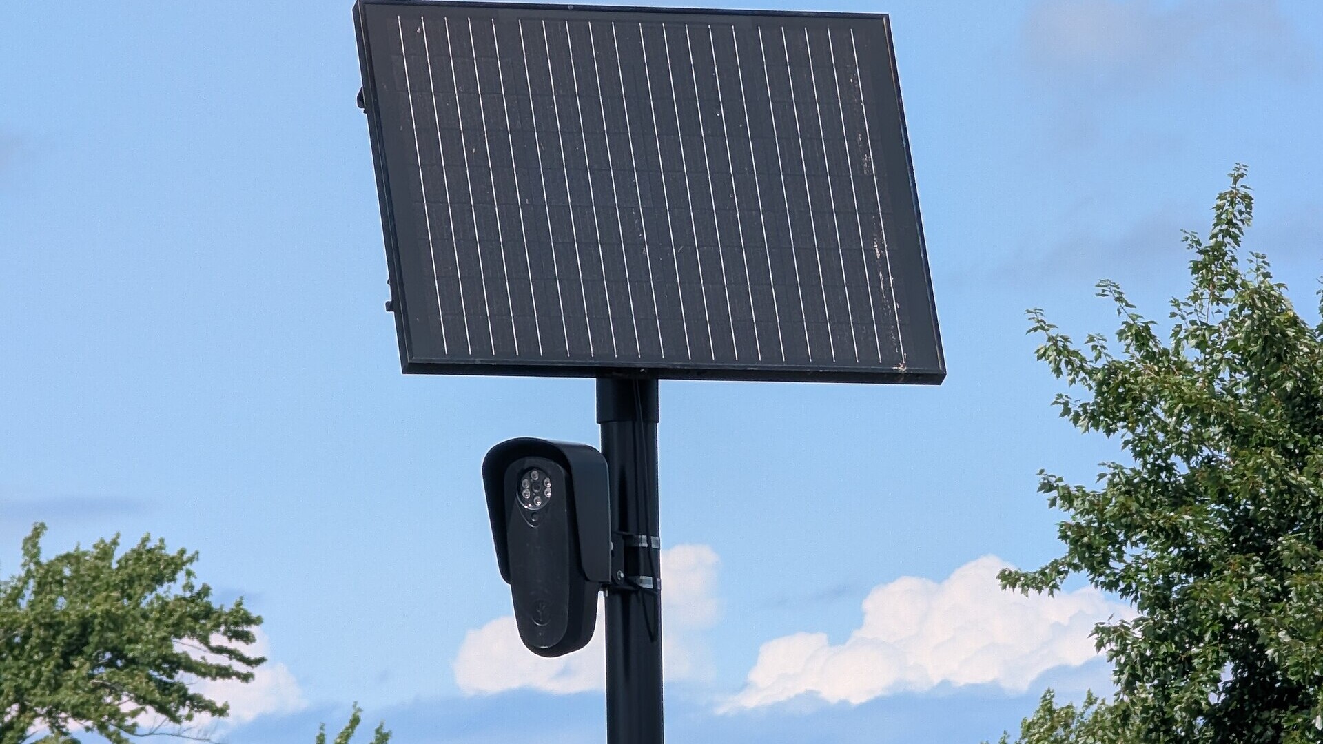 A black Flock Safety license plate reader camera mounted on a pole under a solar panel against a blue sky