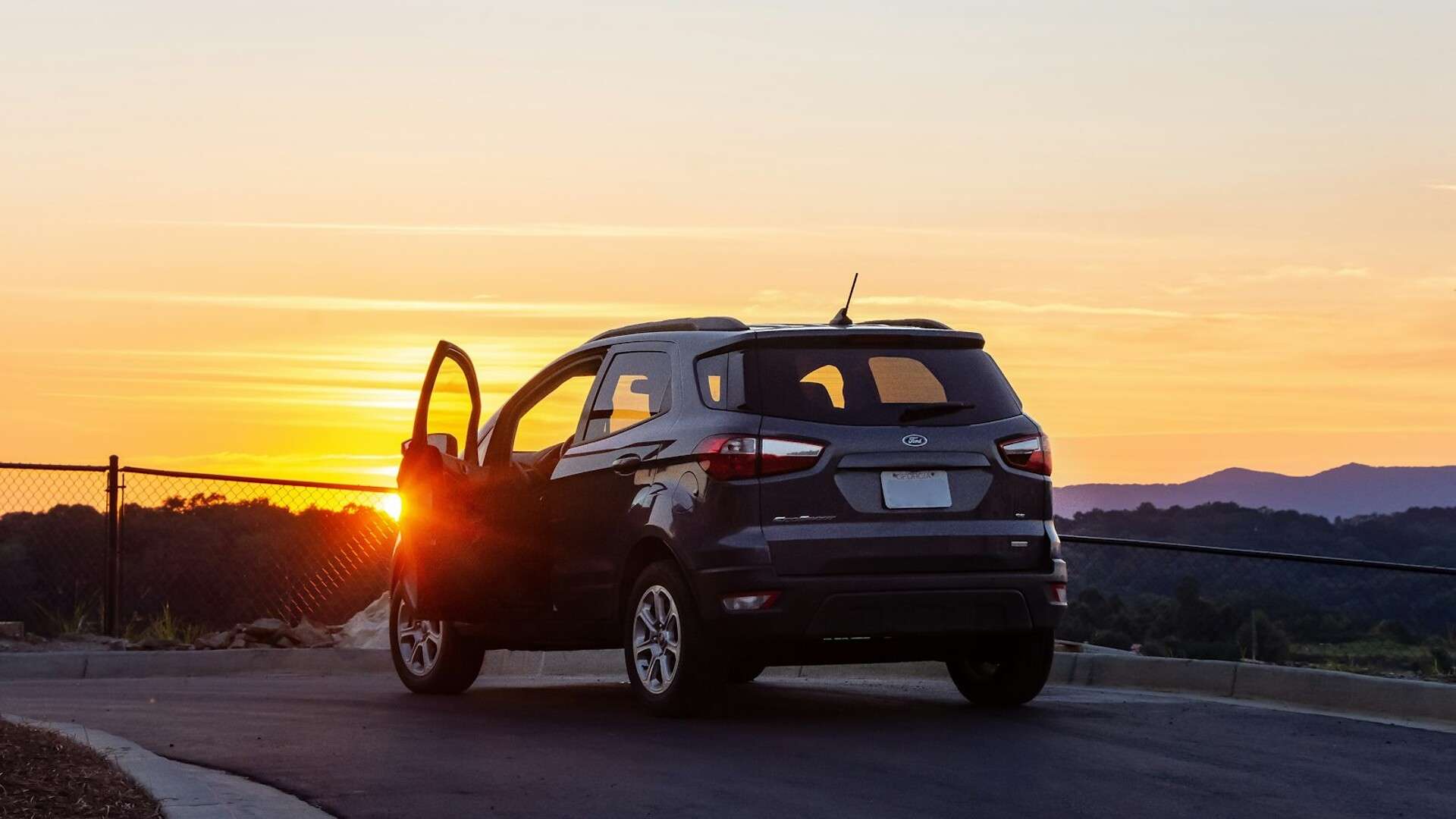 A dark gray Ford EcoSport parked on a hilltop road at sunset with its driver door open