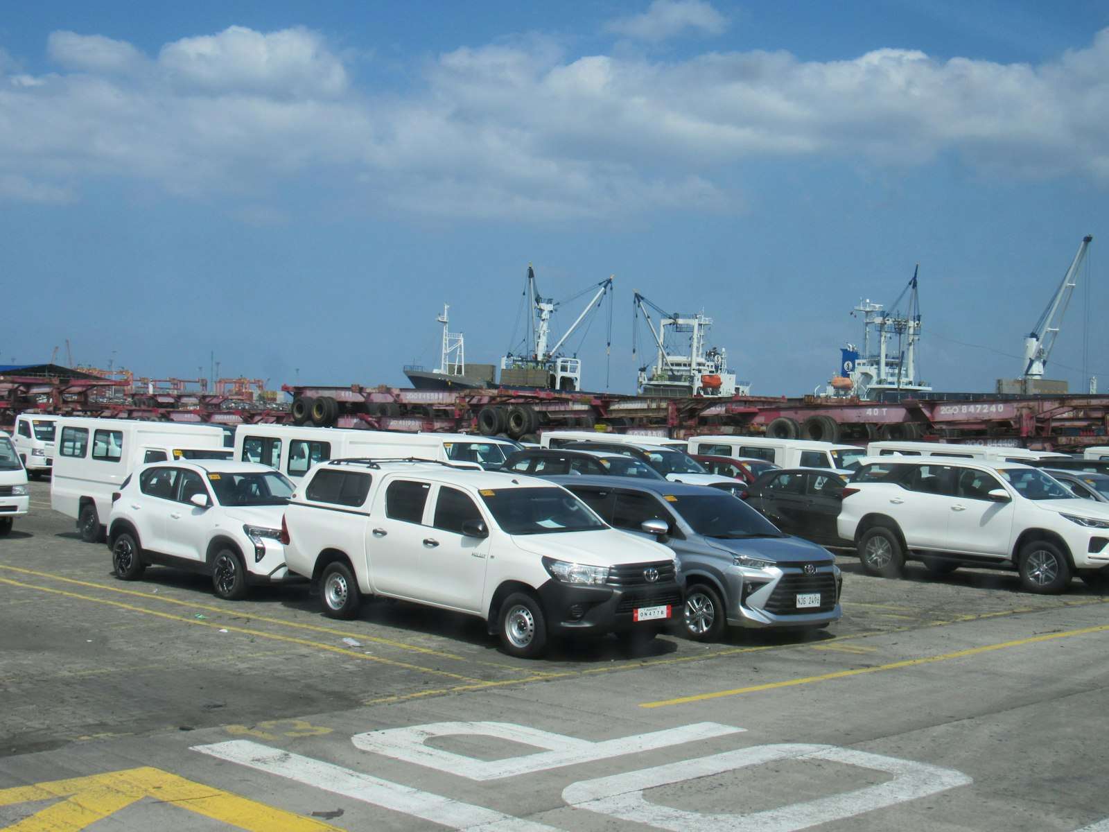Rows of newly imported cars and pickups parked at a port vehicle terminal with cargo ships and cranes behind them