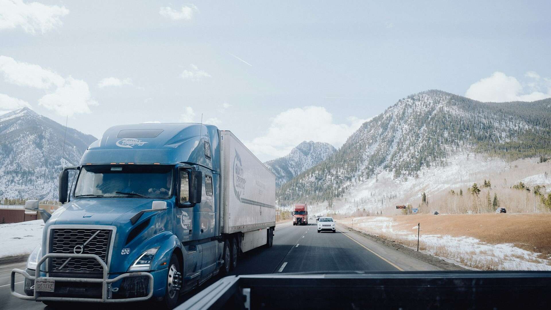 A blue semi truck descends a mountain interstate with snow-covered peaks behind it