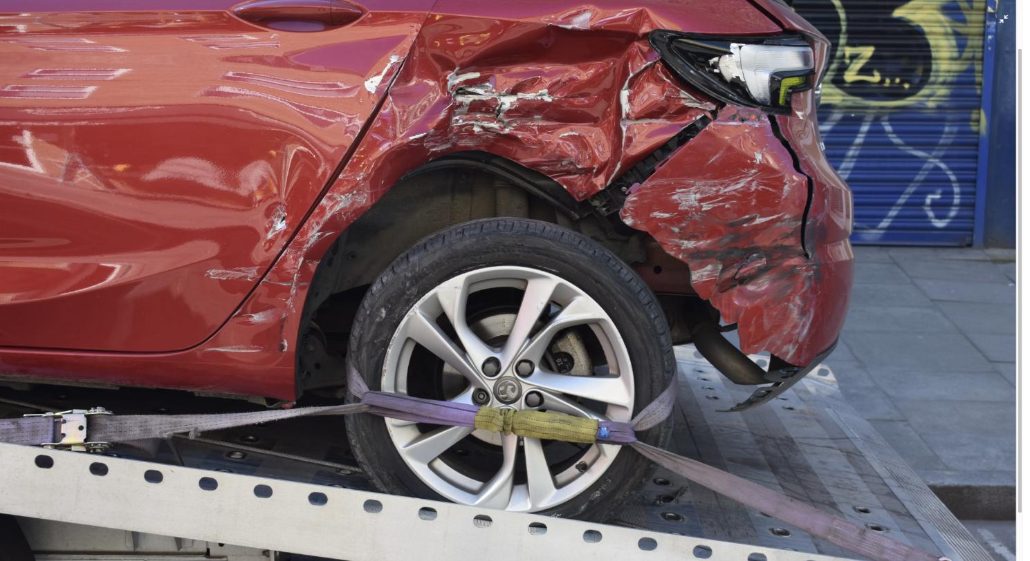 A red car with heavy front-end crash damage strapped to a flatbed tow truck