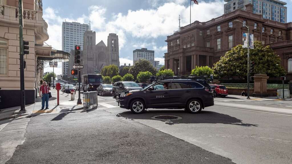 A dark Zoox-branded test SUV with roof-mounted sensors driving through an intersection on California Street in San Francisco