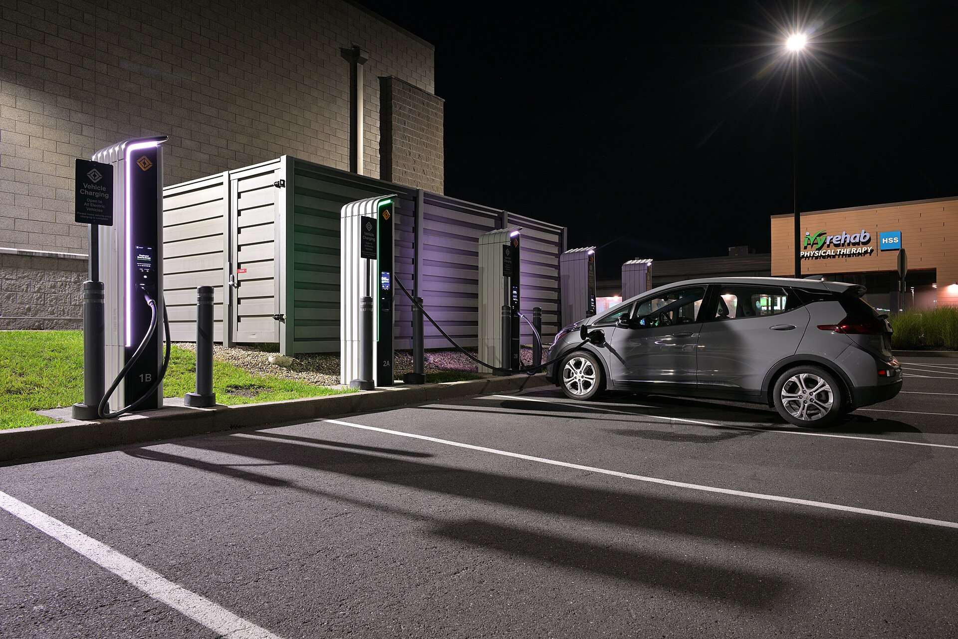 A gray Chevrolet Bolt EV plugged into a DC fast charger at night beside a row of charging cabinets