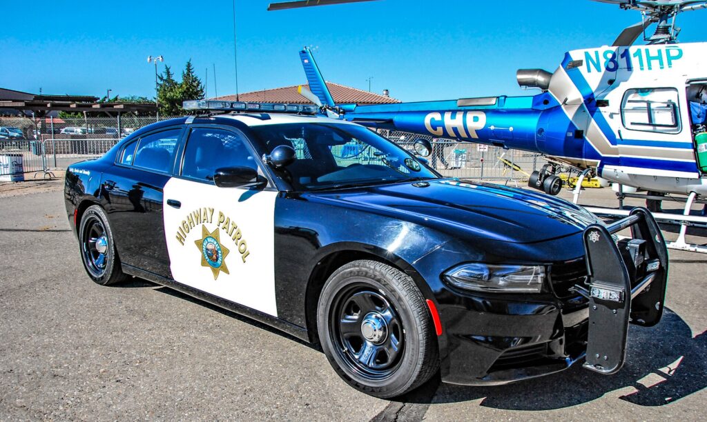 Black-and-white California Highway Patrol Dodge Charger patrol car parked at an outdoor display, with a CHP helicopter behind it