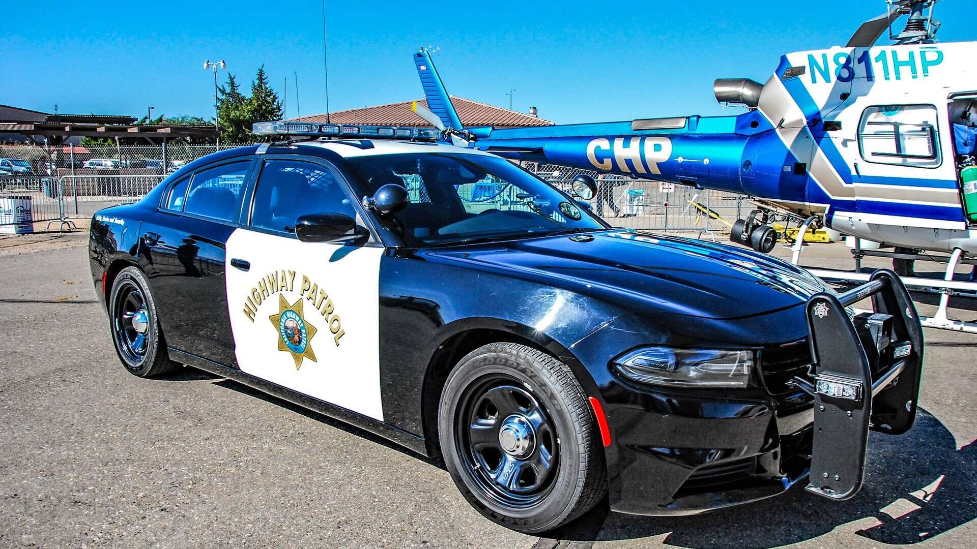 Black-and-white California Highway Patrol Dodge Charger patrol car parked at an outdoor display, with a CHP helicopter behind it