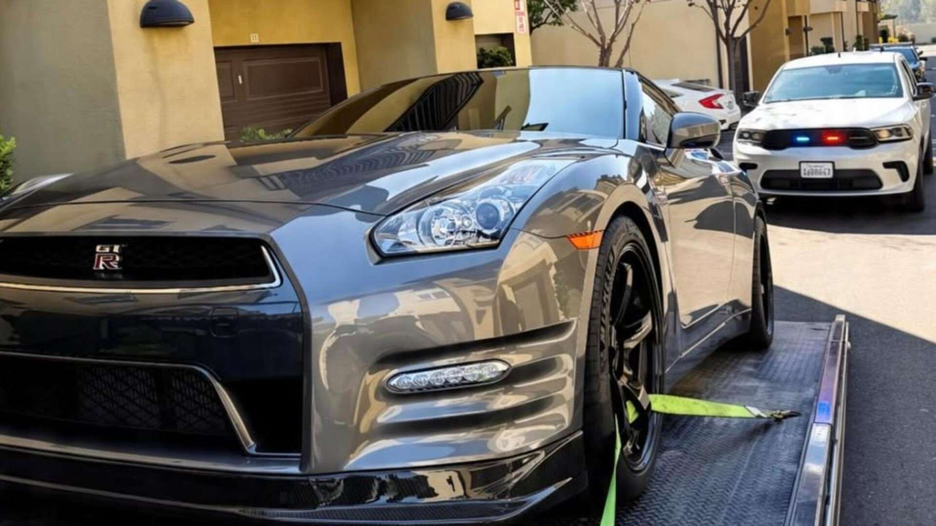 A gray Nissan GT-R strapped to a flatbed tow truck outside a residential building, with a white police SUV behind it