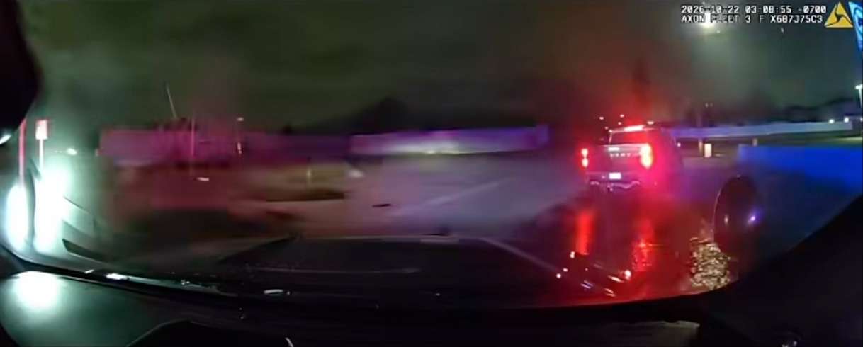 In-car camera view at night of a pickup truck with taillights lit ahead of a patrol vehicle, emergency lights flashing
