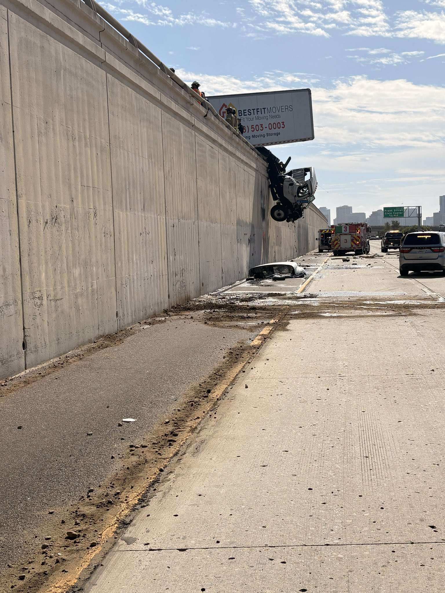 View from the lower I-5 roadway of a truck cab dangling from the top of a tall concrete wall, with debris and spilled material on the pavement below.