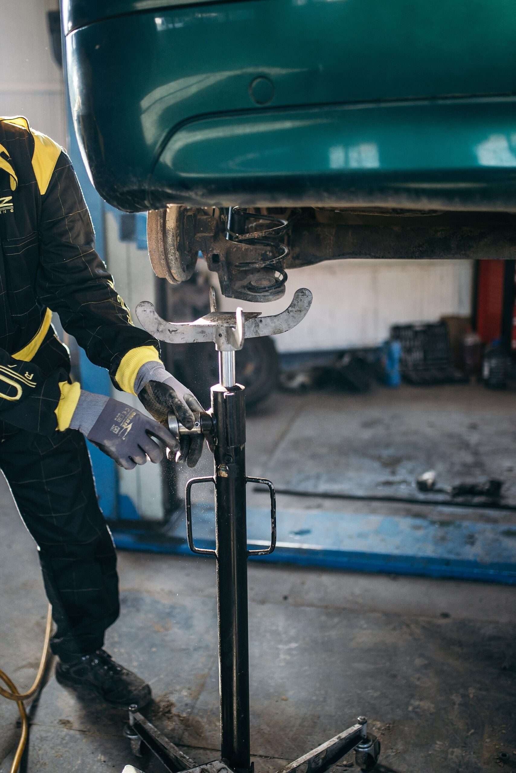A mechanic in gloves adjusts a support stand beneath the coil spring and suspension of a car raised on a lift