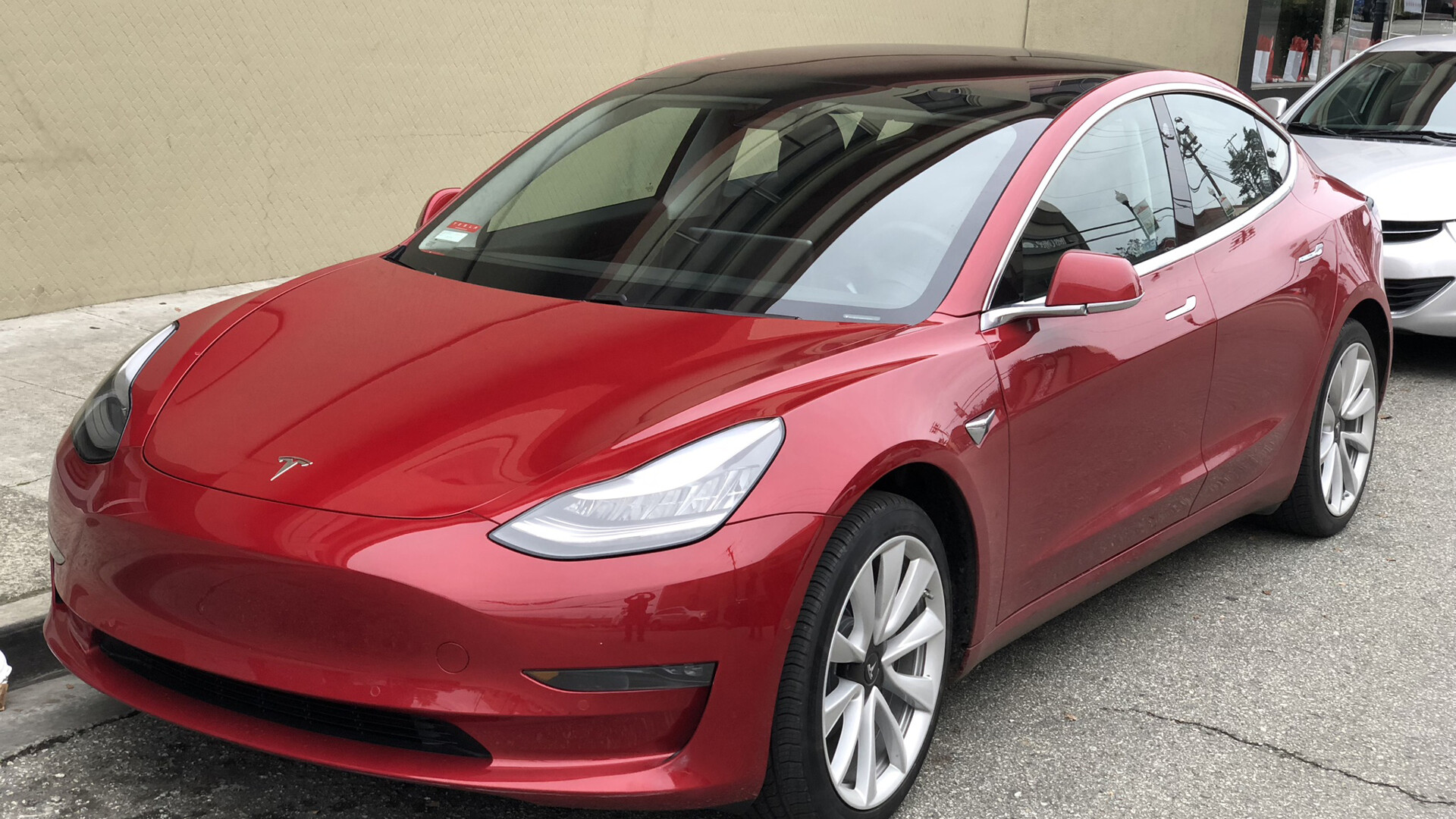 Red first-generation Tesla Model 3 sedan parked at a curb, seen from the front driver side