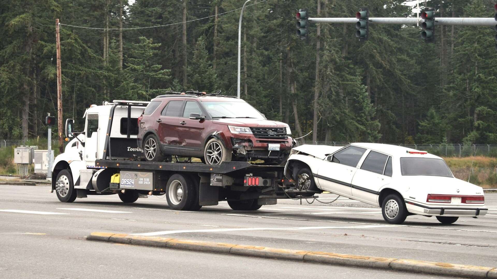 A flatbed tow truck carrying an SUV while towing a white sedan behind it