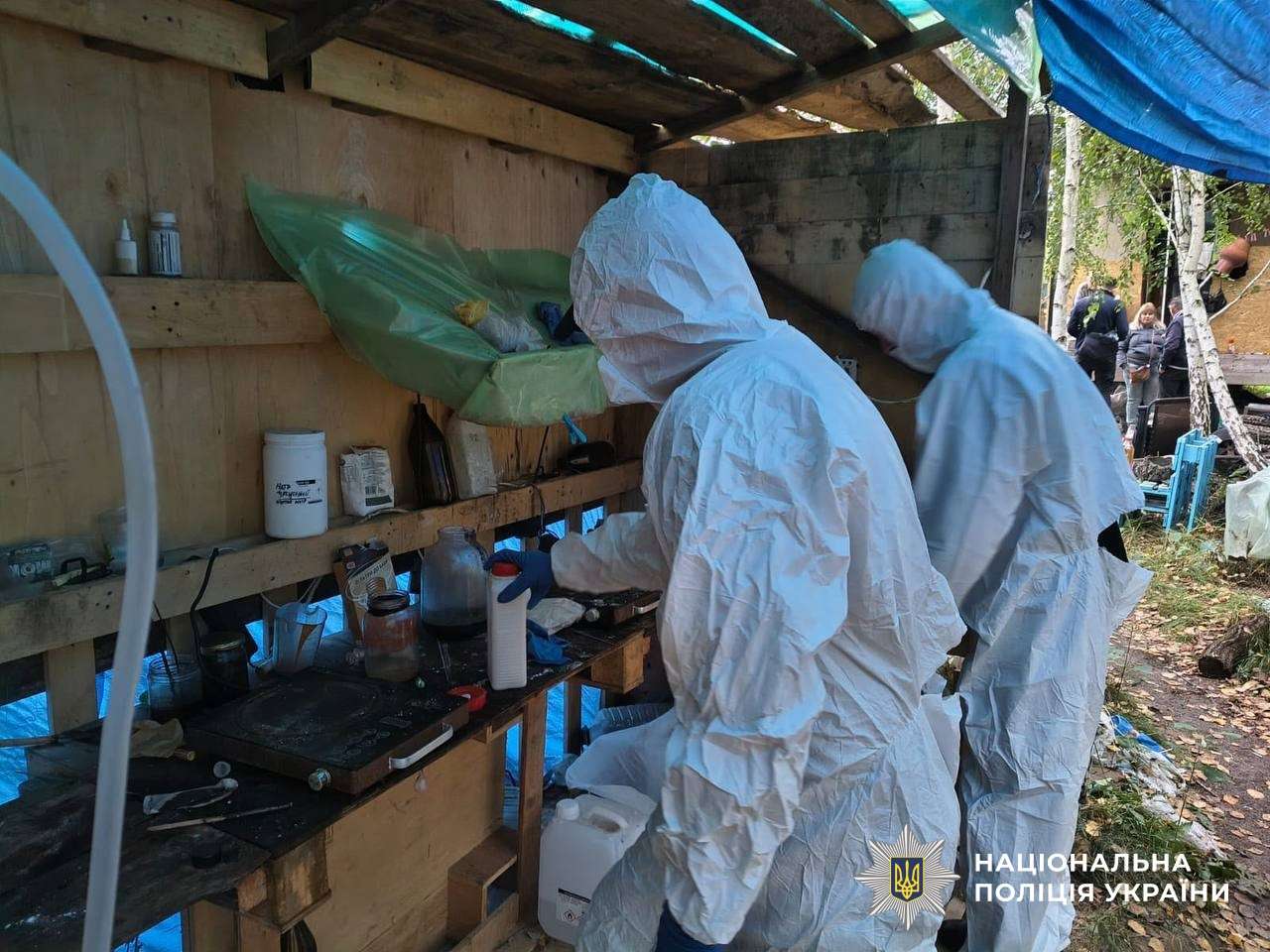 Investigators in protective suits inspect a makeshift outdoor drug lab under a tarp