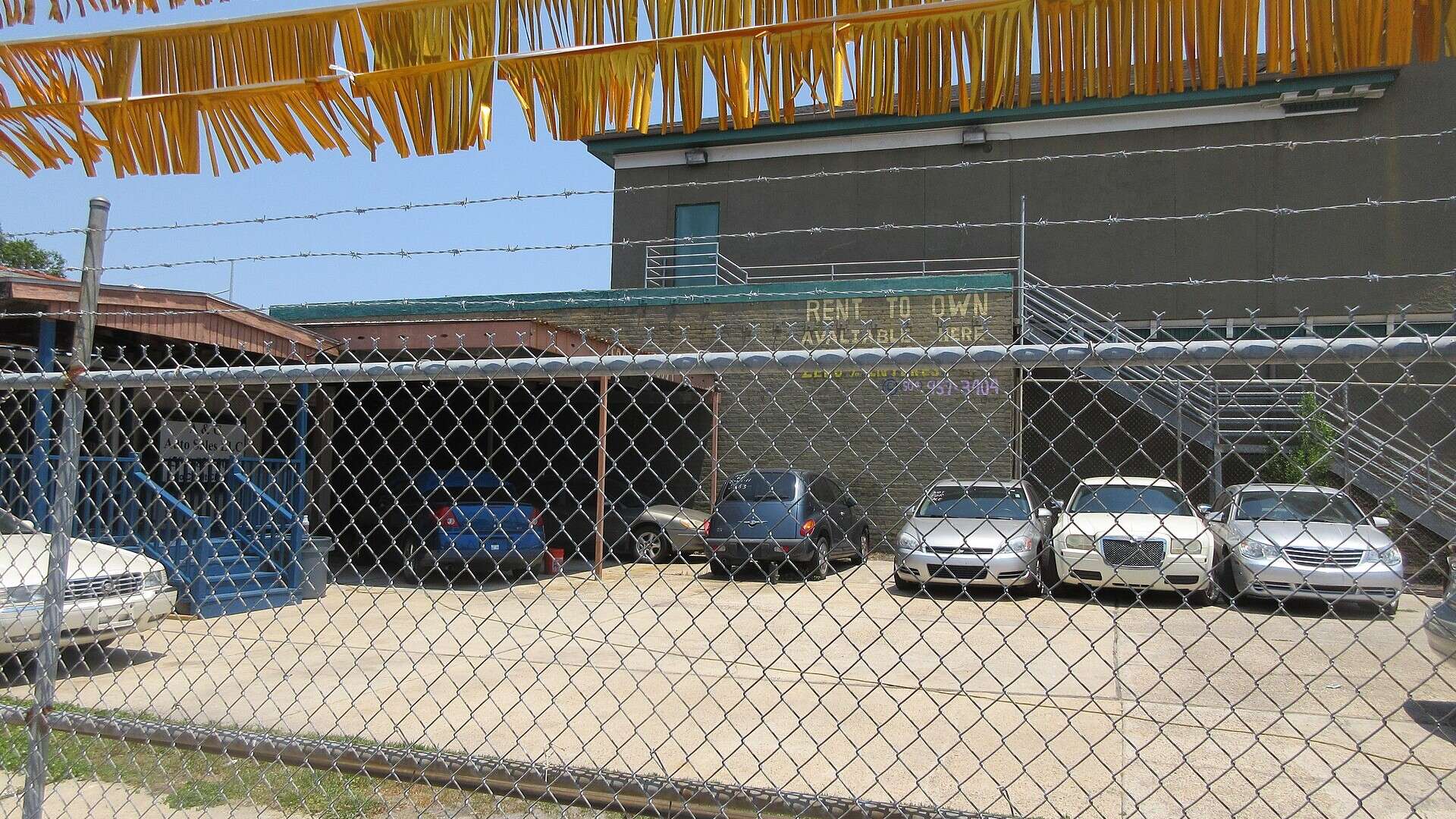 Rows of used sedans parked behind a chain-link fence under strings of orange pennants at a small used car lot