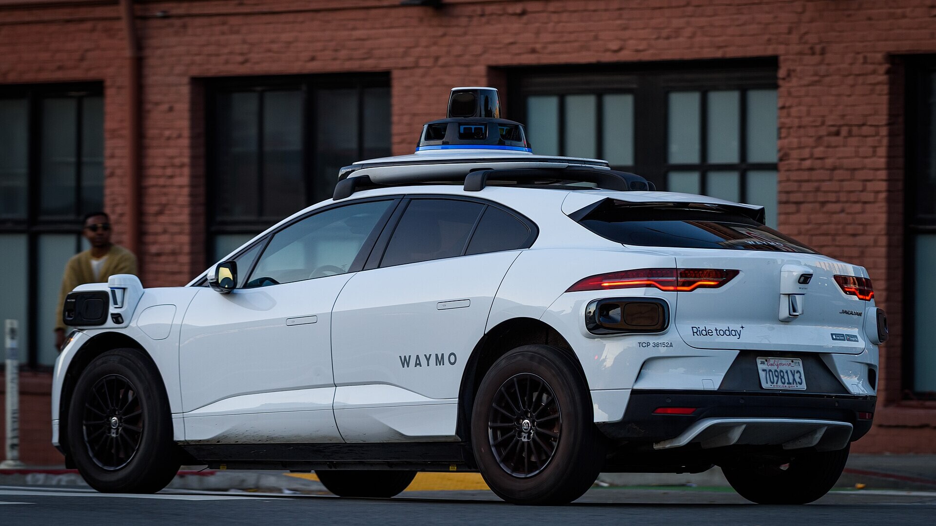 A white Waymo Jaguar I-Pace robotaxi with roof-mounted sensors, seen from the rear quarter