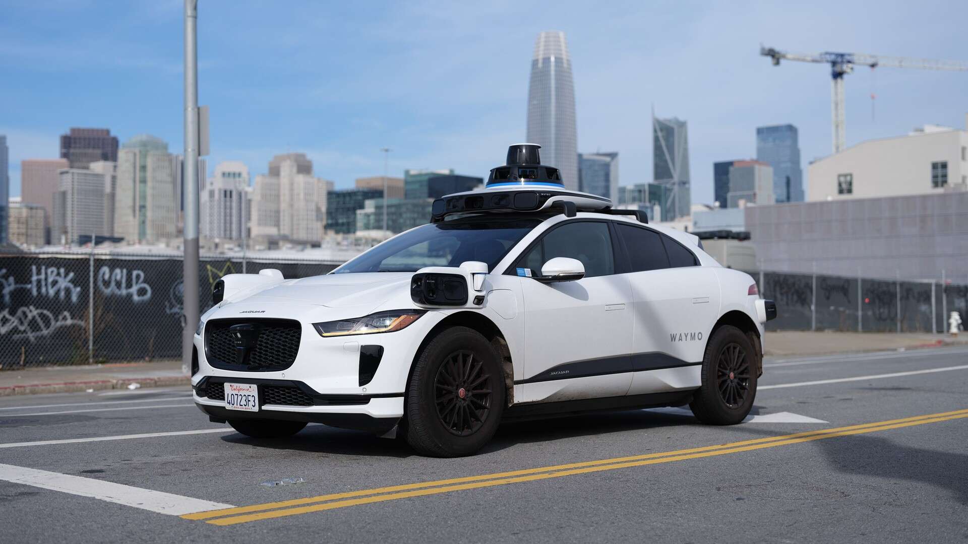 A white Waymo Jaguar I-Pace robotaxi with roof-mounted sensors on a street in San Francisco, with the city skyline behind it