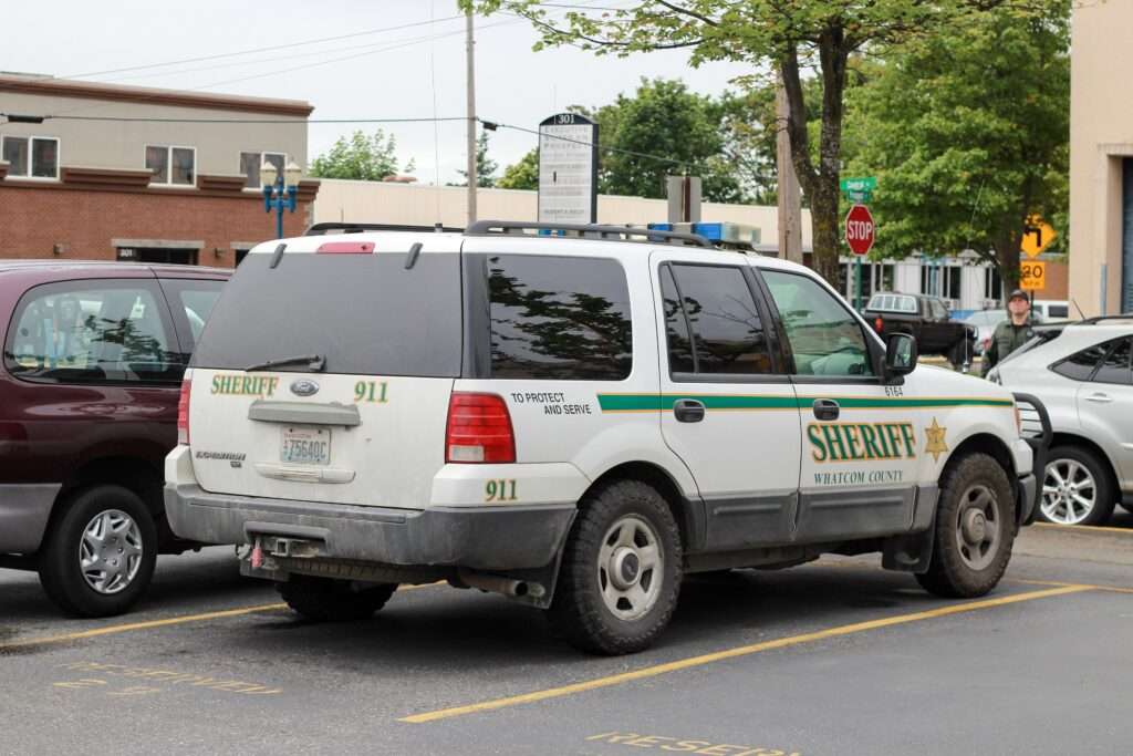 An older white Whatcom County Sheriff SUV with green striping parked on a city street, seen from the rear quarter